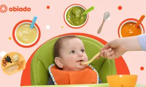 Uśmiechnięte roczne dziecko w krzesełku dostaje jedzenie z łyżeczki. Wokół miseczki z puree i talerzyk z owocami.