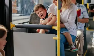 Płaczliwe 1,5 roczne dziecko na rękach mamy w autobusie. Obok siedzi starszy pan z książką.