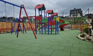 Kolorowy plac zabaw z zieloną, gumową nawierzchnią, huśtawką i zjeżdżalnią.
