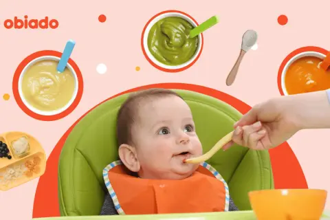 Uśmiechnięte roczne dziecko w krzesełku dostaje jedzenie z łyżeczki. Wokół miseczki z puree i talerzyk z owocami.