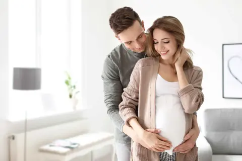 Mężczyzna obejmuje brzuch swojej ciężarnej żony, okazując troskę i miłość. To piękny obraz opieki nad żoną po porodzie, pełen nadziei na przyszłość.