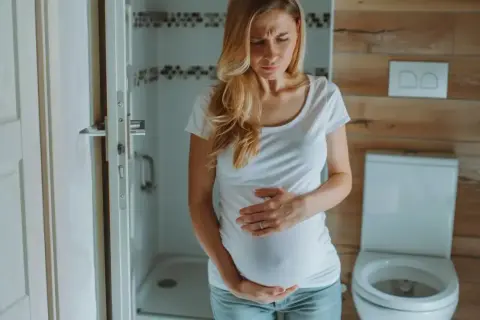 Kobieta w ciąży z wyrazem bólu na twarzy trzyma się za brzuch. W łazience odczuwa ból brzucha w ciąży.