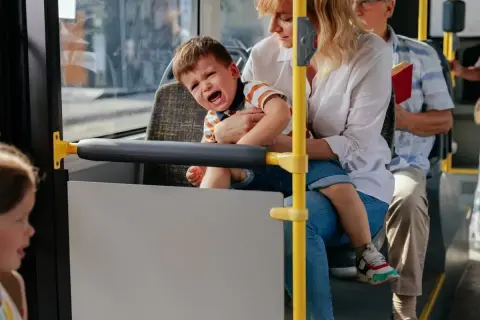 Płaczliwe 1,5 roczne dziecko na rękach mamy w autobusie. Obok siedzi starszy pan z książką.