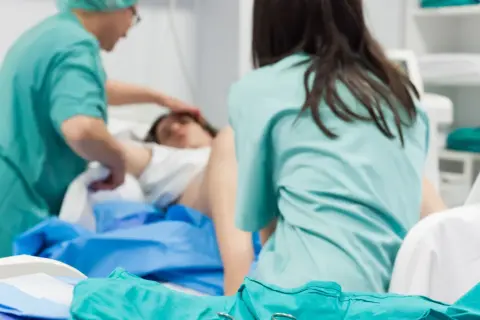 Personel medyczny zajmuje się pacjentką po porodzie. Widoczne są zielone fartuchy i sprzęt medyczny.