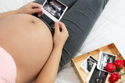 Kiedy widać brzuch ciążowy, przyszła mama trzyma zdjęcie USG. Obok leży pudełko z kolejnymi zdjęciami i czerwonymi różami.