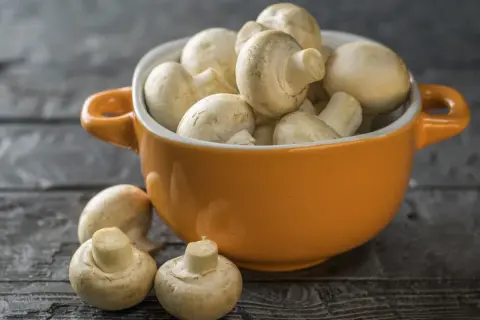 Pomarańczowa miseczka pełna świeżych pieczarek, jakby w ciąży z nowymi smakami. Kilka pieczarek leży obok na drewnianym blacie.