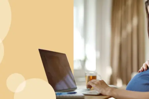 Kobieta w ciąży przy laptopie, która uśmiecha się, dotykając brzucha. Dowiaduje się, jak liczyć ruchy dziecka.