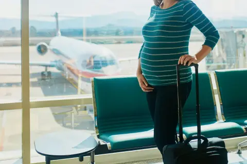 Czekając na lotnisku, przyszła mama zastanawia się, czy w ciąży można latać samolotem. Obok walizka, przed nią samolot.