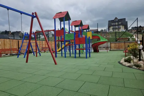 Kolorowy plac zabaw z zieloną, gumową nawierzchnią, huśtawką i zjeżdżalnią.