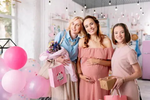 Trzy uśmiechnięte kobiety świętują baby shower po polsku. W tle balony i prezenty.