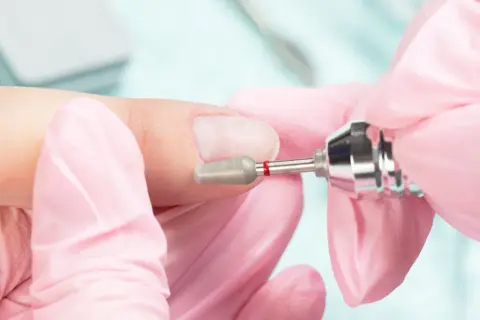 Dłonie w różowych rękawiczkach wykonują manicure frezarką. Przygotowanie paznokci do porodu, by były schludne i bezpieczne.