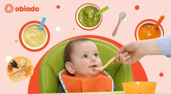 Uśmiechnięte roczne dziecko w krzesełku dostaje jedzenie z łyżeczki. Wokół miseczki z puree i talerzyk z owocami.