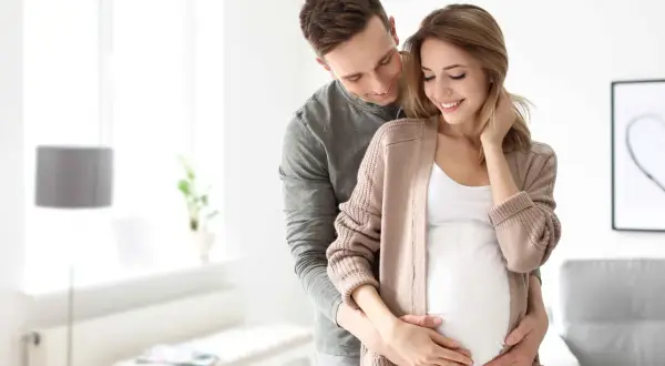 Mężczyzna obejmuje brzuch swojej ciężarnej żony, okazując troskę i miłość. To piękny obraz opieki nad żoną po porodzie, pełen nadziei na przyszłość.