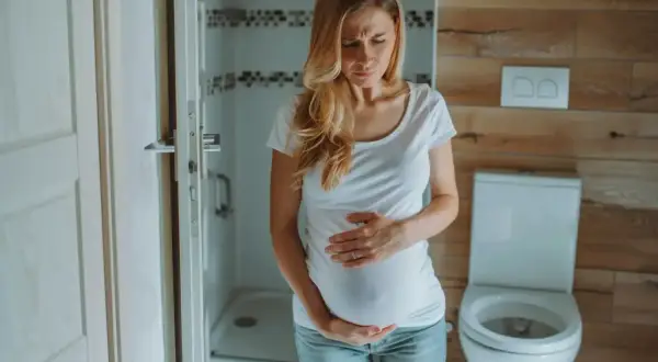 Kobieta w ciąży z wyrazem bólu na twarzy trzyma się za brzuch. W łazience odczuwa ból brzucha w ciąży.