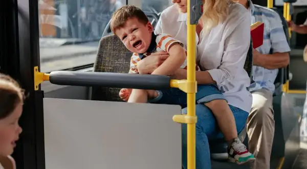 Płaczliwe 1,5 roczne dziecko na rękach mamy w autobusie. Obok siedzi starszy pan z książką.