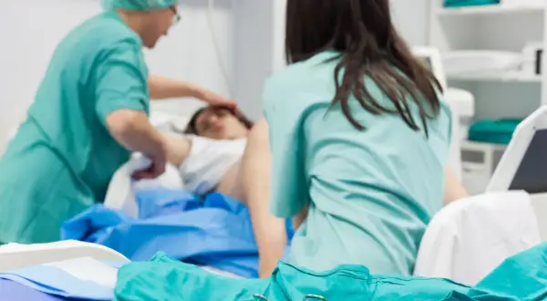 Personel medyczny zajmuje się pacjentką po porodzie. Widoczne są zielone fartuchy i sprzęt medyczny.