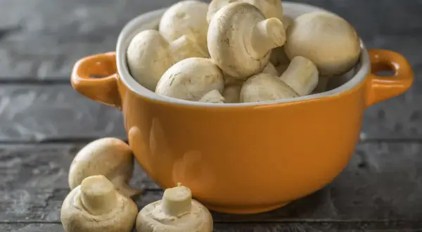 Pomarańczowa miseczka pełna świeżych pieczarek, jakby w ciąży z nowymi smakami. Kilka pieczarek leży obok na drewnianym blacie.