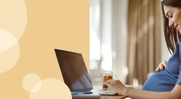 Kobieta w ciąży przy laptopie, która uśmiecha się, dotykając brzucha. Dowiaduje się, jak liczyć ruchy dziecka.