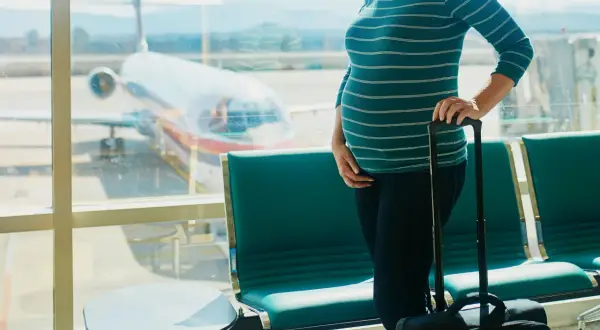 Czekając na lotnisku, przyszła mama zastanawia się, czy w ciąży można latać samolotem. Obok walizka, przed nią samolot.