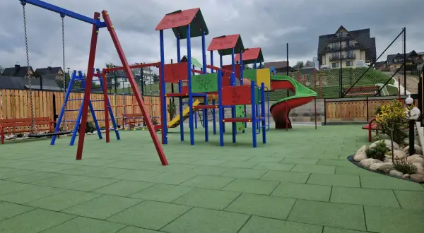 Kolorowy plac zabaw z zieloną, gumową nawierzchnią, huśtawką i zjeżdżalnią.