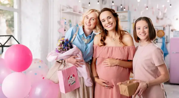 Trzy uśmiechnięte kobiety świętują baby shower po polsku. W tle balony i prezenty.