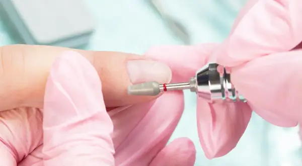 Dłonie w różowych rękawiczkach wykonują manicure frezarką. Przygotowanie paznokci do porodu, by były schludne i bezpieczne.