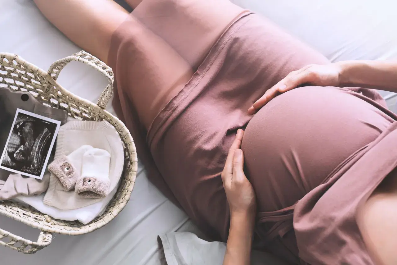 Brzuszek ciężarnej kobiety, na którym spoczywają dłonie. Obok koszyk z pierwszymi ubrankami, skarpetkami i zdjęciem USG, które pokazuje maleństwo w worku owodniowym.