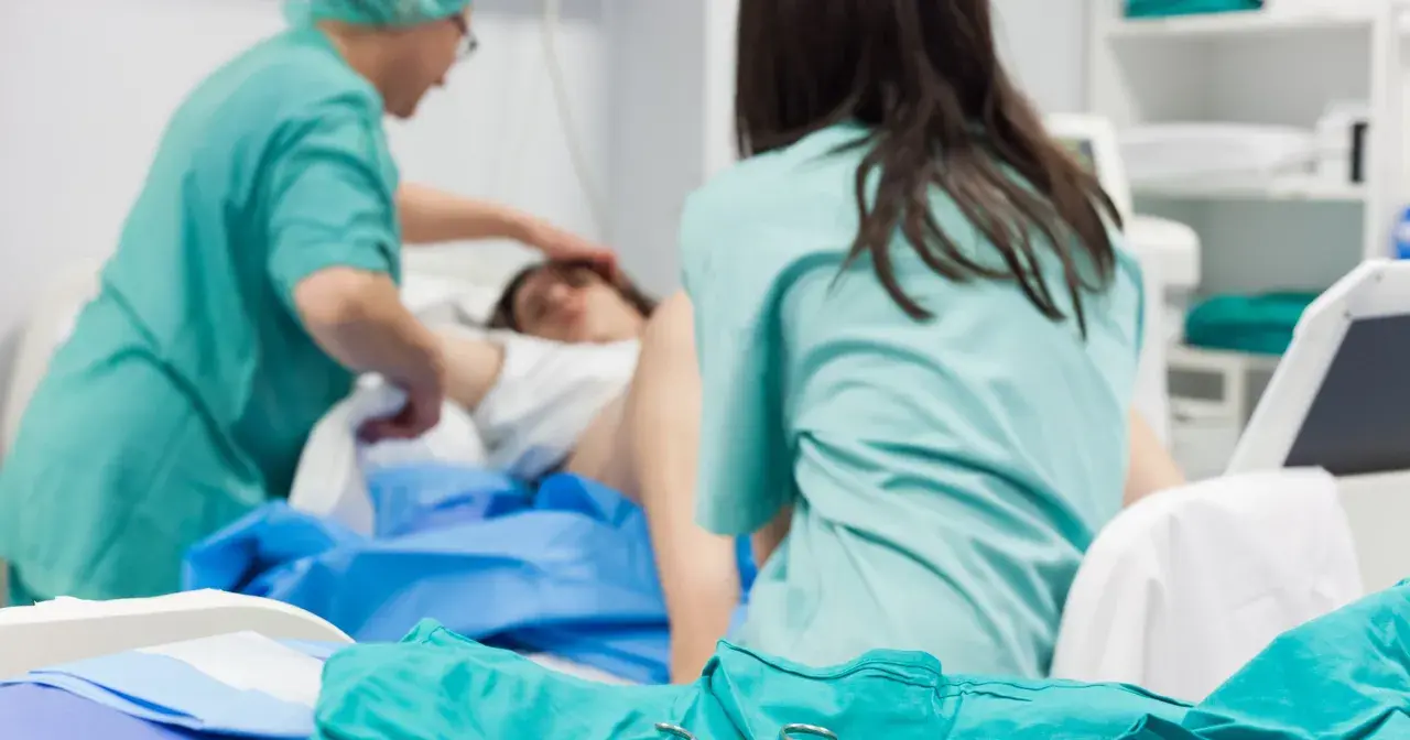 Personel medyczny zajmuje się pacjentką po porodzie. Widoczne są zielone fartuchy i sprzęt medyczny.