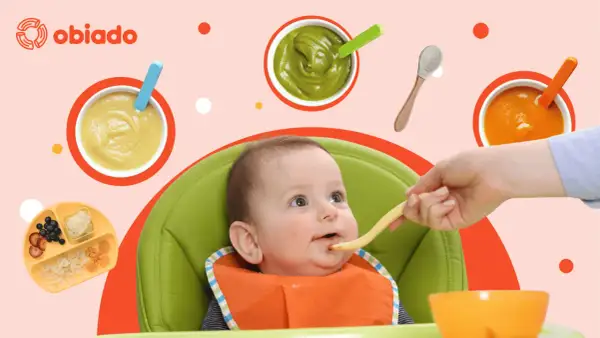 Uśmiechnięte roczne dziecko w krzesełku dostaje jedzenie z łyżeczki. Wokół miseczki z puree i talerzyk z owocami.