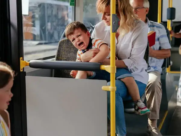 Płaczliwe 1,5 roczne dziecko na rękach mamy w autobusie. Obok siedzi starszy pan z książką.